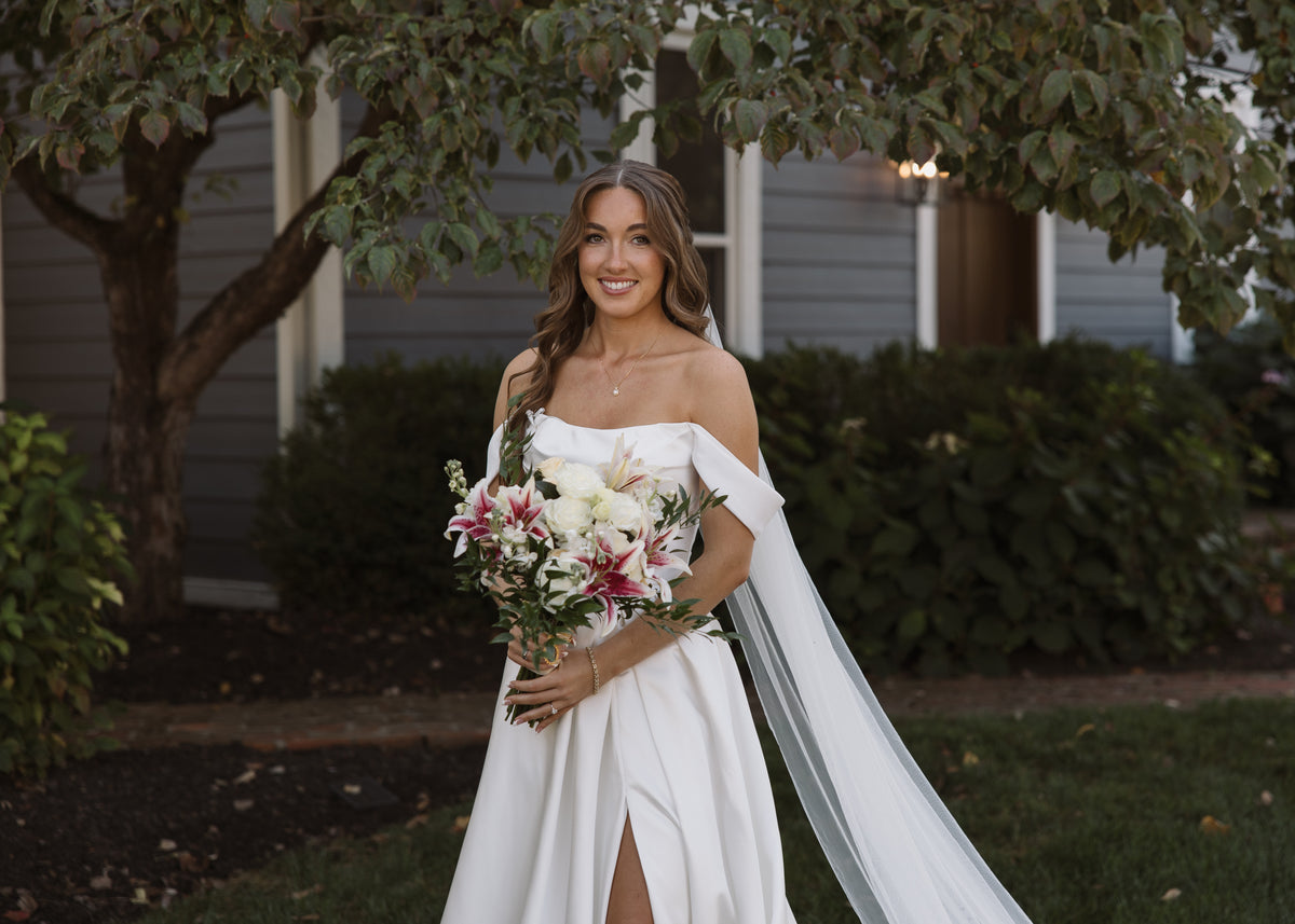 Bride holding a white bouquet with pink-centered lilies and greenery beneath a leafy tree.