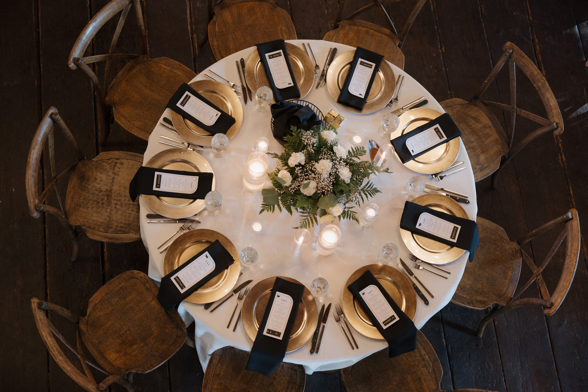 Overhead view of a round reception table with a white floral centerpiece, candles, gold chargers and dark napkins.