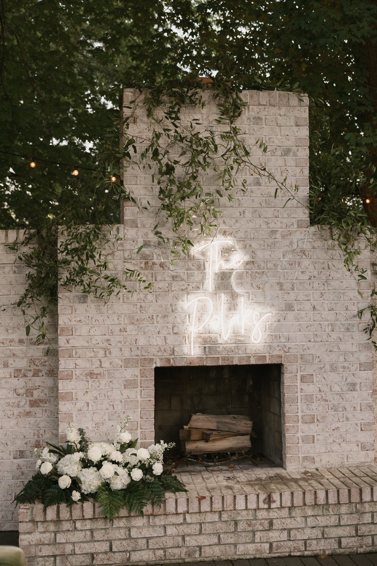 White flowers at the base of an outdoor brick fireplace, with trailing greenery and an illuminated wedding sign above.