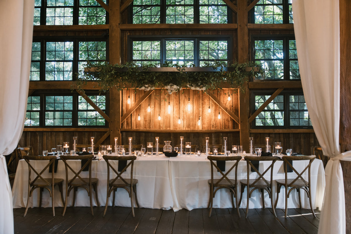 Long reception table beneath suspended greenery in a timber room with tall windows and white drapes.