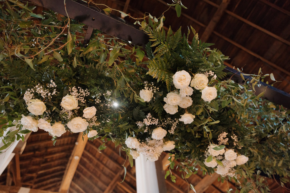 White flowers and layered greenery suspended from an overhead ladder beneath timber roof beams.
